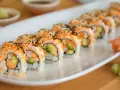 Rząd roladek sushi california roll, posypanych sezamem i ikrą, z awokado i łososiem w środku. Obok imbir marynowany i ogórek.