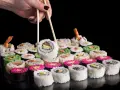 Dłoń z pałeczkami podnosi sushi. Różnorodne rolki, w tym zielone i różowe, czekają na spróbowanie. Nauka jak jeść pałeczkami to klucz do tej uczty.