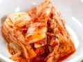 Domowe kimchi w białej misce, z widocznymi liśćmi kapusty i przyprawami.