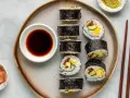Na talerzu ułożone są sushi: kilka mniejszych hosomak i większe futomaki, obok miseczka sosu sojowego i marynowanego imbiru.