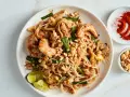 Talerz z Pad Thai z krewetkami, orzeszkami ziemnymi, jajkiem i limonką, obok miseczki z orzeszkami i papryką.