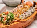 Drewniana łódka pełna sushi, dania z kraju, z którego pochodzi sushi, czyli Japonii. Obok stos białych talerzy.