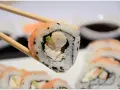 Chopsticks trzymają sushi z krewetkami, awokado i kremowym serkiem. W tle widać więcej roladek sushi.