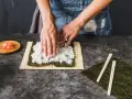 Dłonie rozprowadzają ryż na nori, przygotowując się do zwinięcia sushi. Obok leżą pałeczki i marynowany imbir, idealny dodatek do sushi.