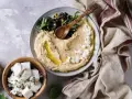Kremowy hummus z ciecierzycy, posypany fetą i oliwkami, z drewnianą łyżką. Obok miseczka z kostkami sera feta.
