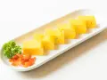 Sześć kawałków tamago, czyli japońskiego omletu, ułożonych na białej desce. Obok natka pietruszki i pomarańczowe dodatki.