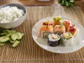 Kolorowe rolki sushi bez nori z łososiem, awokado i innymi dodatkami. Obok miska ryżu i pokrojony ogórek.