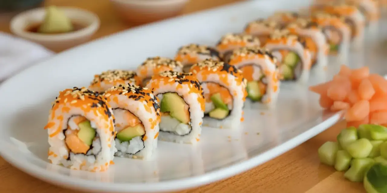 Rząd roladek sushi california roll, posypanych sezamem i ikrą, z awokado i łososiem w środku. Obok imbir marynowany i ogórek.