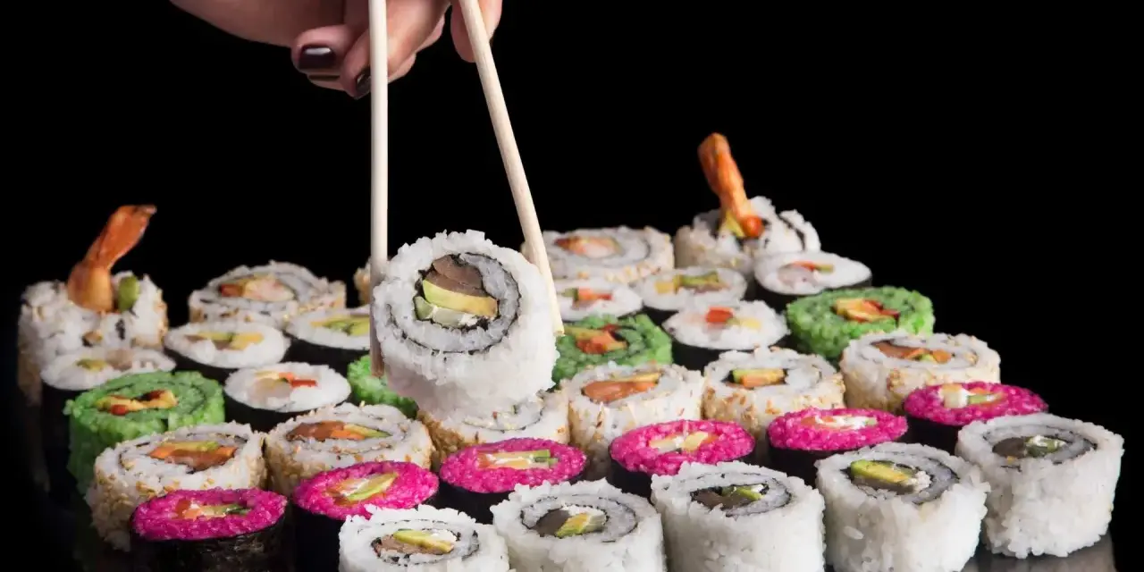 Dłoń z pałeczkami podnosi sushi. Różnorodne rolki, w tym zielone i różowe, czekają na spróbowanie. Nauka jak jeść pałeczkami to klucz do tej uczty.
