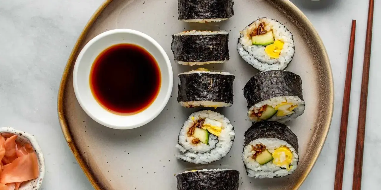 Na talerzu ułożone są sushi: kilka mniejszych hosomak i większe futomaki, obok miseczka sosu sojowego i marynowanego imbiru.