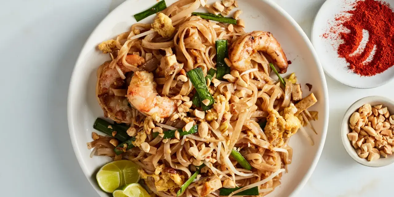 Talerz z Pad Thai z krewetkami, orzeszkami ziemnymi, jajkiem i limonką, obok miseczki z orzeszkami i papryką.