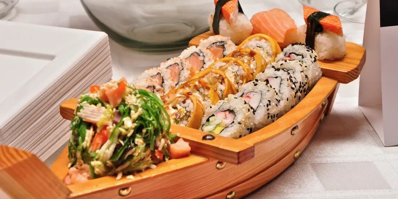Drewniana łódka pełna sushi, dania z kraju, z którego pochodzi sushi, czyli Japonii. Obok stos białych talerzy.