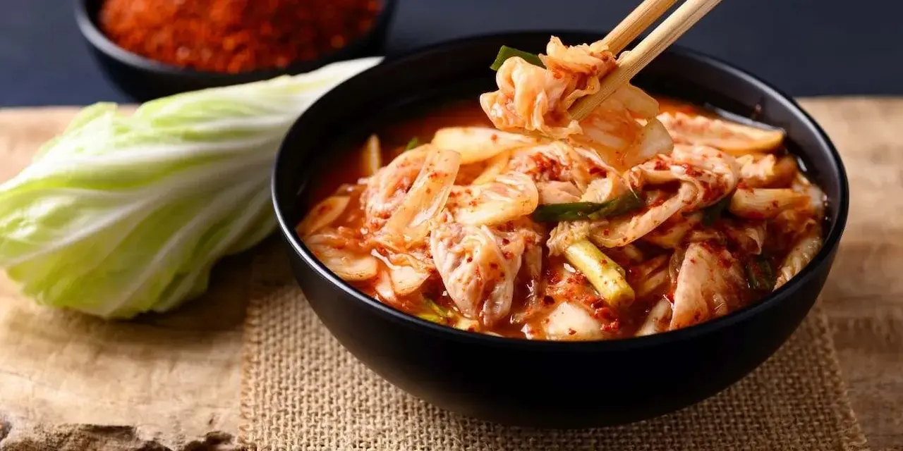 Miseczka z kimchi, pałeczki podnoszące kawałek, kapusta i chili w tle.