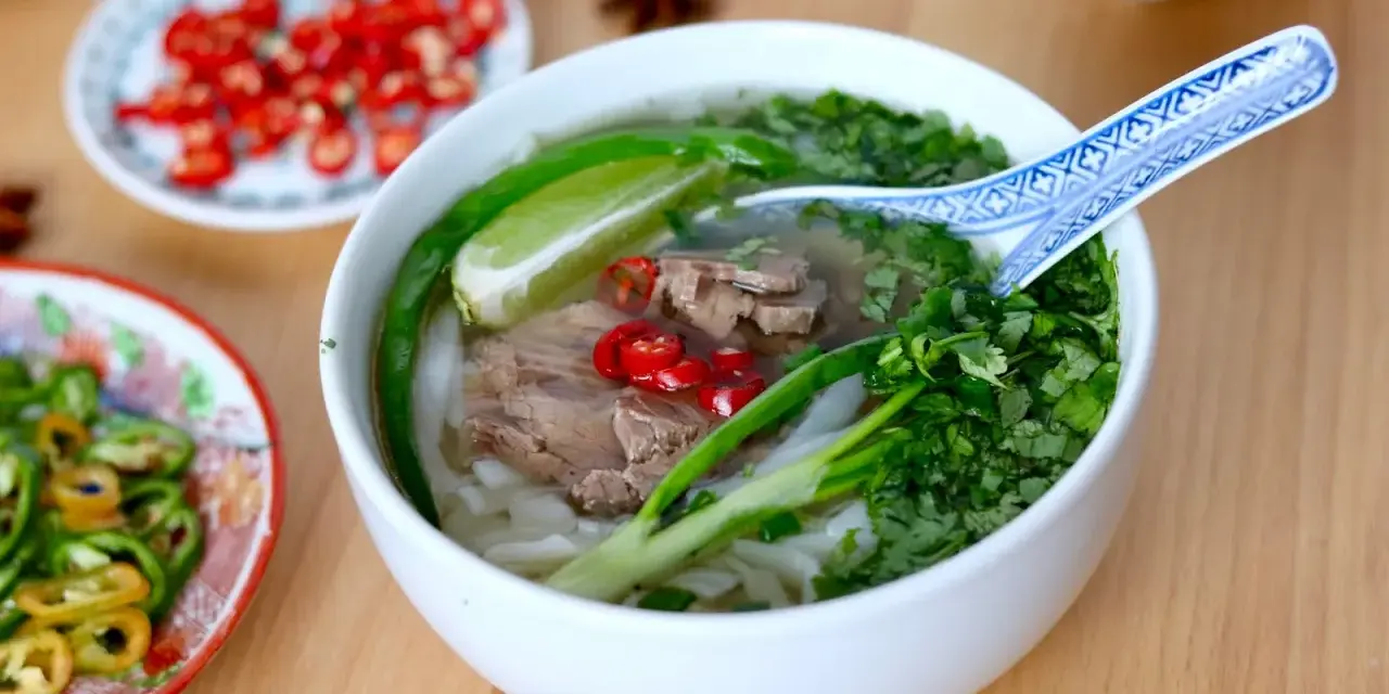 Aromatyczna zupa pho z wołowiną, makaronem ryżowym, świeżą kolendrą, chili i limonką. Obok talerzyki z dodatkami.