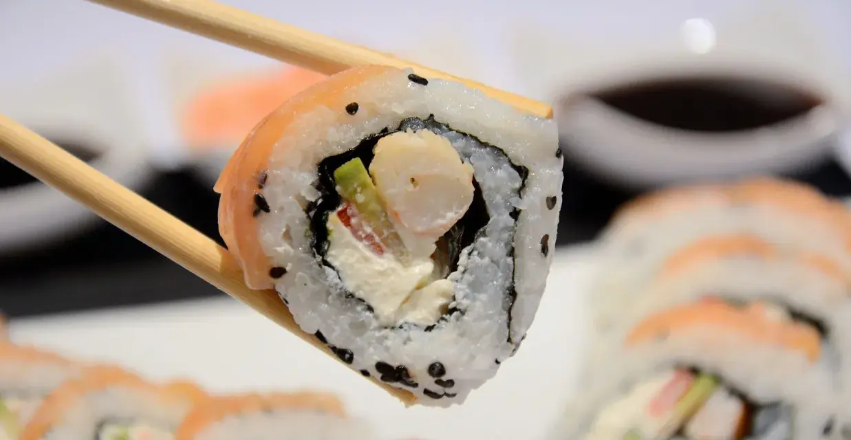 Chopsticks trzymają sushi z krewetkami, awokado i kremowym serkiem. W tle widać więcej roladek sushi.