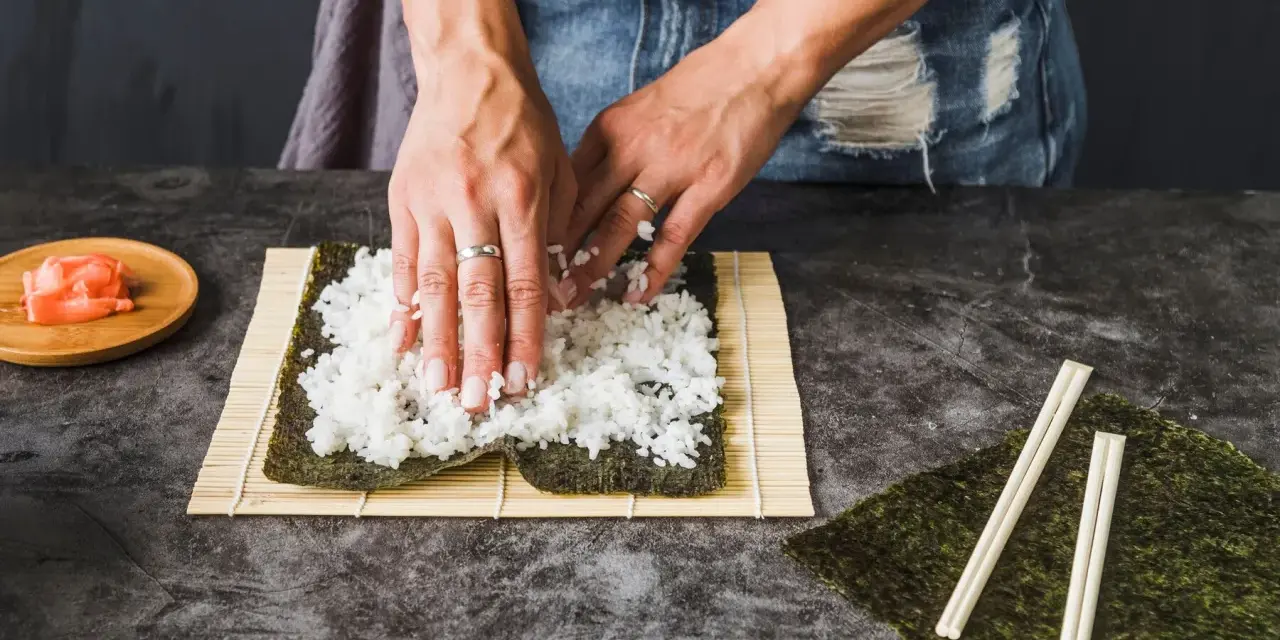 Dłonie rozprowadzają ryż na nori, przygotowując się do zwinięcia sushi. Obok leżą pałeczki i marynowany imbir, idealny dodatek do sushi.