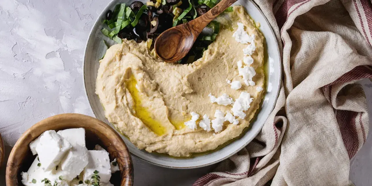 Kremowy hummus z ciecierzycy, posypany fetą i oliwkami, z drewnianą łyżką. Obok miseczka z kostkami sera feta.