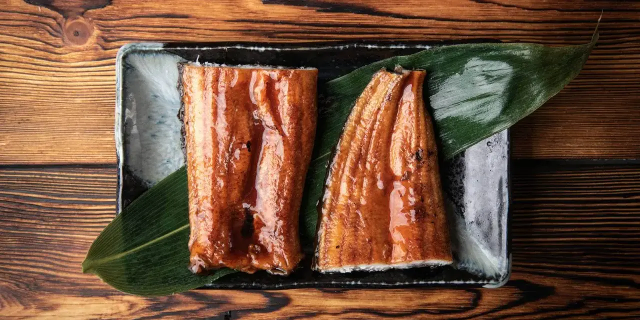 Dwa kawałki błyszczącego, glazurowanego sushi z węgorzem na liściu, podane na czarnej tacce.