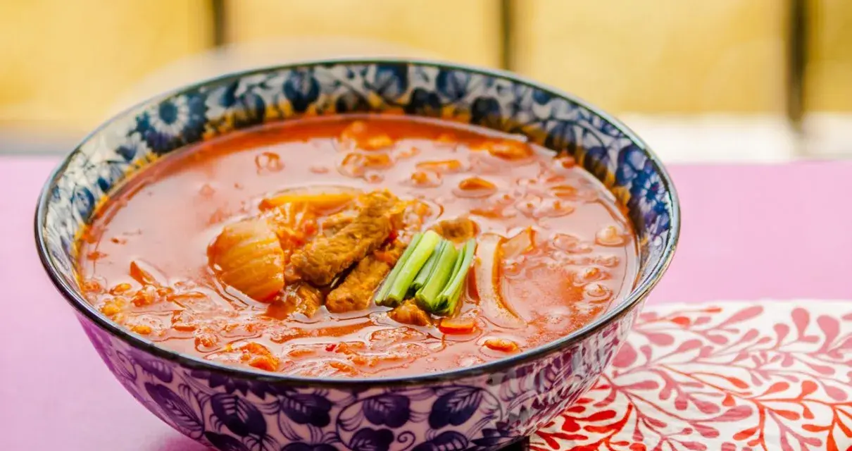 Gorąca zupa z kimchi z kawałkami mięsa i zieloną cebulką, podana w ozdobnej misce.