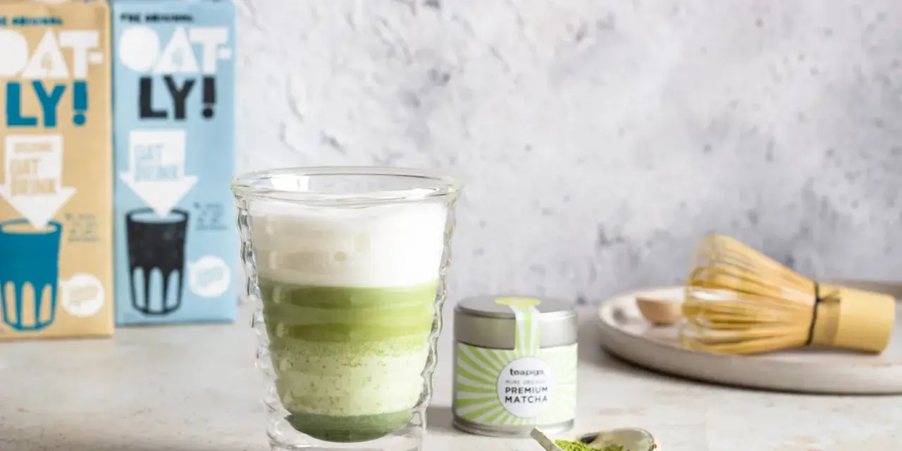 Orzeźwiający iced matcha latte z pianką, obok puszka matcha i bambusowa miotełka. Idealny napój na ciepłe dni.