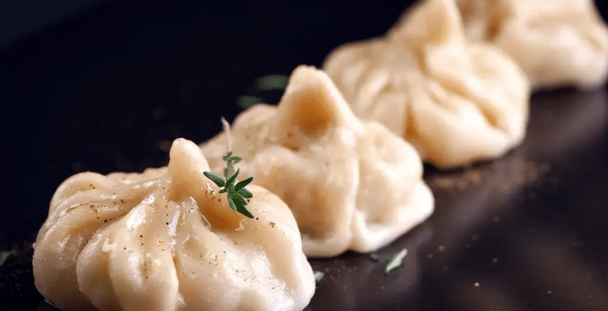 A row of delicate pierożki z krewetkami, garnished with fresh herbs and spices, presented on a dark plate.