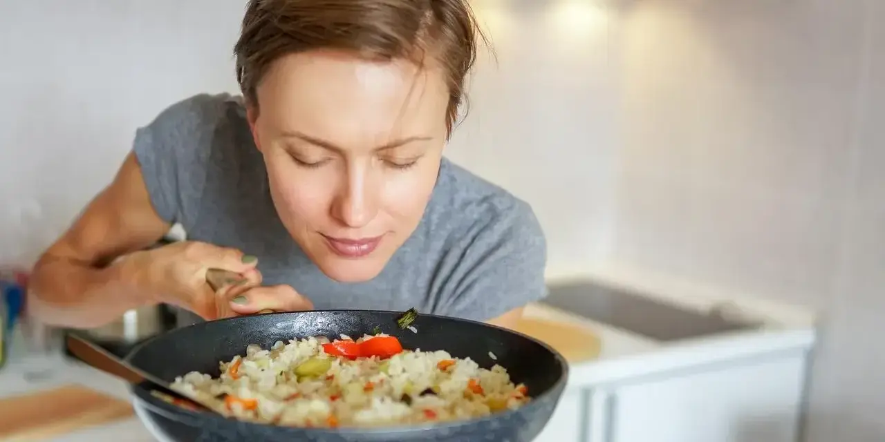 Kobieta wącha ryż z warzywami na patelni, zastanawiając się, jak przyprawić ryż, by był idealny.