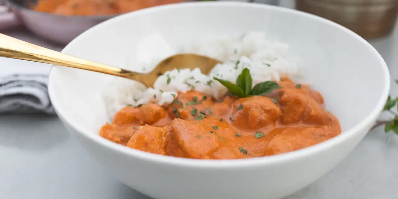 Aksamitny butter chicken masala z ryżem, udekorowany miętą. Złota łyżka zanurzona w daniu.