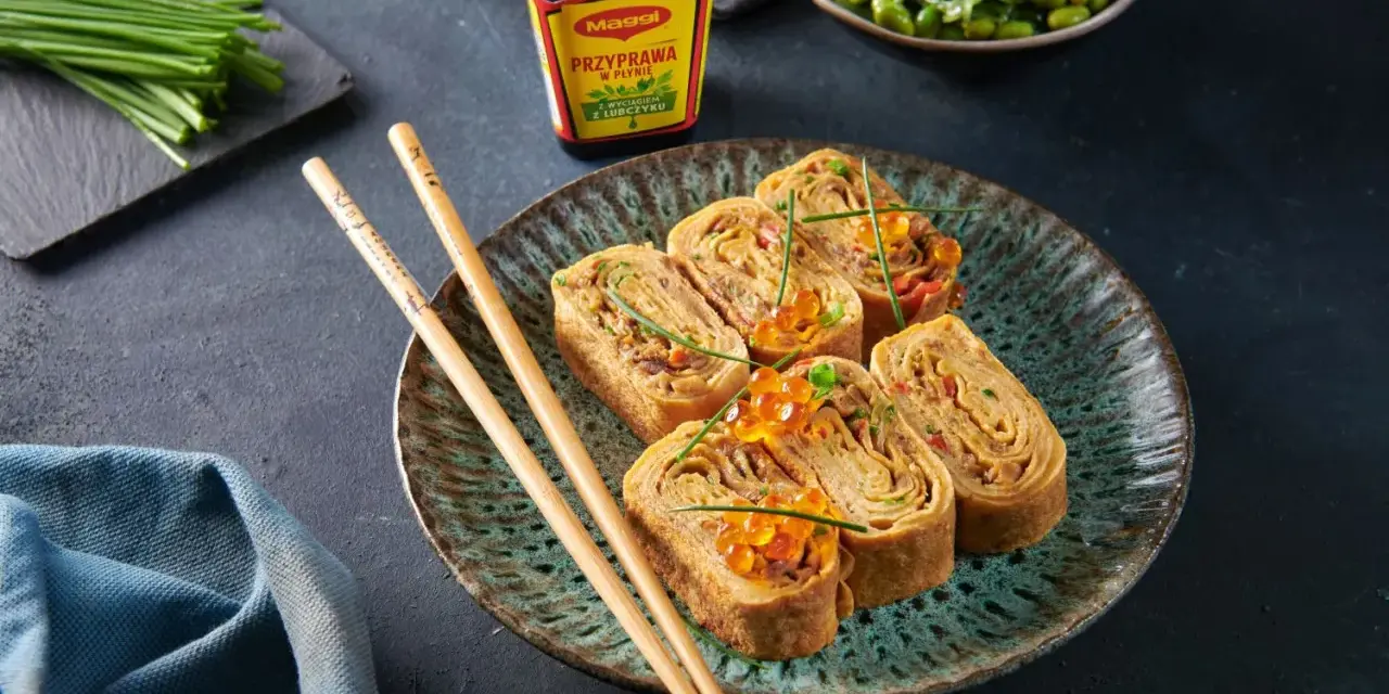 Roladki z omletu do sushi, ozdobione ikrą łososia i szczypiorkiem, obok pałeczki i sosu Maggi.