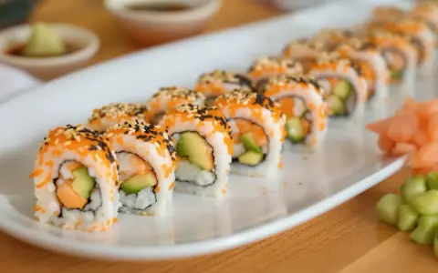 Rząd roladek sushi california roll, posypanych sezamem i ikrą, z awokado i łososiem w środku. Obok imbir marynowany i ogórek.