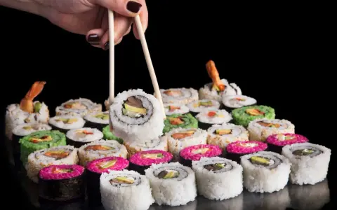 Dłoń z pałeczkami podnosi sushi. Różnorodne rolki, w tym zielone i różowe, czekają na spróbowanie. Nauka jak jeść pałeczkami to klucz do tej uczty.