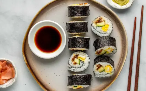 Na talerzu ułożone są sushi: kilka mniejszych hosomak i większe futomaki, obok miseczka sosu sojowego i marynowanego imbiru.