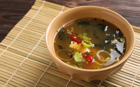 Aksamitna zupa miso z tofu, wodorostami, szczypiorkiem i papryczką chili, podana w ceramicznej miseczce na bambusowej macie.