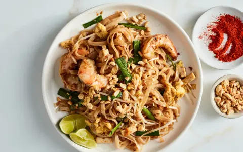Talerz z Pad Thai z krewetkami, orzeszkami ziemnymi, jajkiem i limonką, obok miseczki z orzeszkami i papryką.