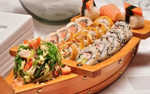 Drewniana łódka pełna sushi, dania z kraju, z którego pochodzi sushi, czyli Japonii. Obok stos białych talerzy.