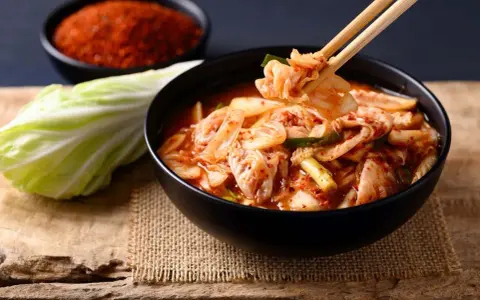 Miseczka z kimchi, pałeczki podnoszące kawałek, kapusta i chili w tle.