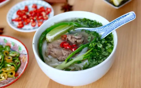 Aromatyczna zupa pho z wołowiną, makaronem ryżowym, świeżą kolendrą, chili i limonką. Obok talerzyki z dodatkami.