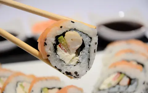 Chopsticks trzymają sushi z krewetkami, awokado i kremowym serkiem. W tle widać więcej roladek sushi.
