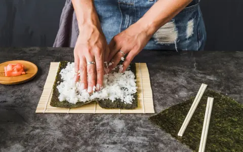 Dłonie rozprowadzają ryż na nori, przygotowując się do zwinięcia sushi. Obok leżą pałeczki i marynowany imbir, idealny dodatek do sushi.