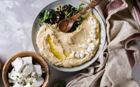 Kremowy hummus z ciecierzycy, posypany fetą i oliwkami, z drewnianą łyżką. Obok miseczka z kostkami sera feta.