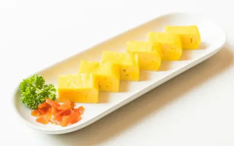 Sześć kawałków tamago, czyli japońskiego omletu, ułożonych na białej desce. Obok natka pietruszki i pomarańczowe dodatki.