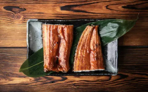 Dwa kawałki błyszczącego, glazurowanego sushi z węgorzem na liściu, podane na czarnej tacce.