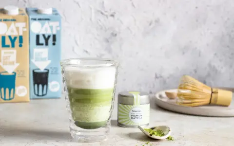 Orzeźwiający iced matcha latte z pianką, obok puszka matcha i bambusowa miotełka. Idealny napój na ciepłe dni.