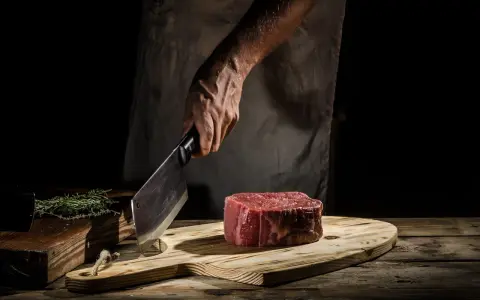 Mistrz kuchni pokazuje, jak kroic wolowine w paski. Na desce leży kawałek surowej wołowiny, obok gałązka rozmarynu.
