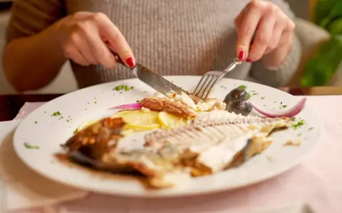Osoba kroi widelcem i nożem rybę, która wygląda jak dorsz w ciąży, z ziemniakami, oliwkami i cebulą.