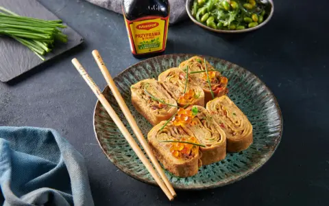 Roladki z omletu do sushi, ozdobione ikrą łososia i szczypiorkiem, obok pałeczki i sosu Maggi.