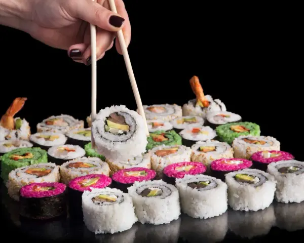 Dłoń z pałeczkami podnosi sushi. Różnorodne rolki, w tym zielone i różowe, czekają na spróbowanie. Nauka jak jeść pałeczkami to klucz do tej uczty.