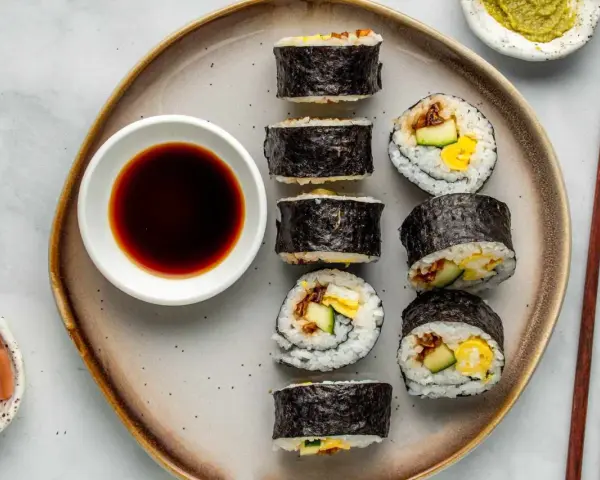 Na talerzu ułożone są sushi: kilka mniejszych hosomak i większe futomaki, obok miseczka sosu sojowego i marynowanego imbiru.