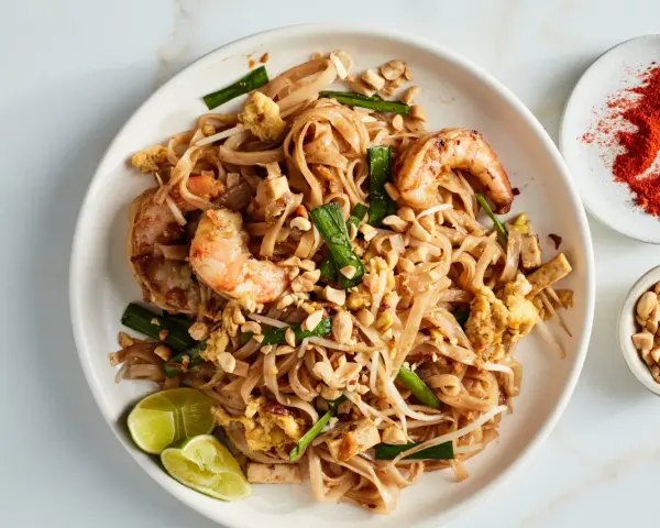 Talerz z Pad Thai z krewetkami, orzeszkami ziemnymi, jajkiem i limonką, obok miseczki z orzeszkami i papryką.