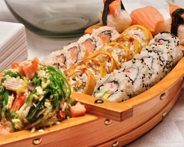 Drewniana łódka pełna sushi, dania z kraju, z którego pochodzi sushi, czyli Japonii. Obok stos białych talerzy.