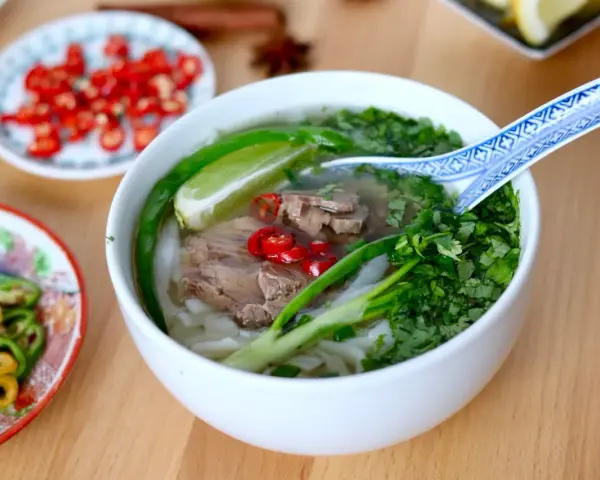 Aromatyczna zupa pho z wołowiną, makaronem ryżowym, świeżą kolendrą, chili i limonką. Obok talerzyki z dodatkami.